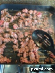 Mouthwatering Slow Cooker Taco Meat 3 Ground beef is sizzling in a grill pan on the stovetop, perfect for a mild chili recipe. A black slotted spatula rests nearby as steam rises from the browning meat. The pan's grill lines sear the beef while bits of onion add flavor, hinting at delicious possibilities ahead.