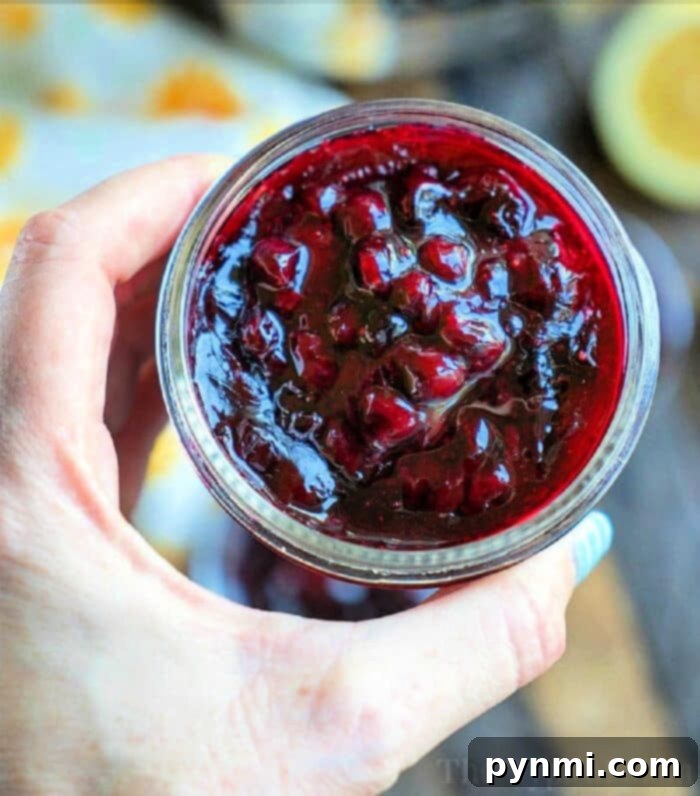 A hand demonstrating how to can jam holds a jar of homemade blackberry jam, showcasing the visible fruit pieces.