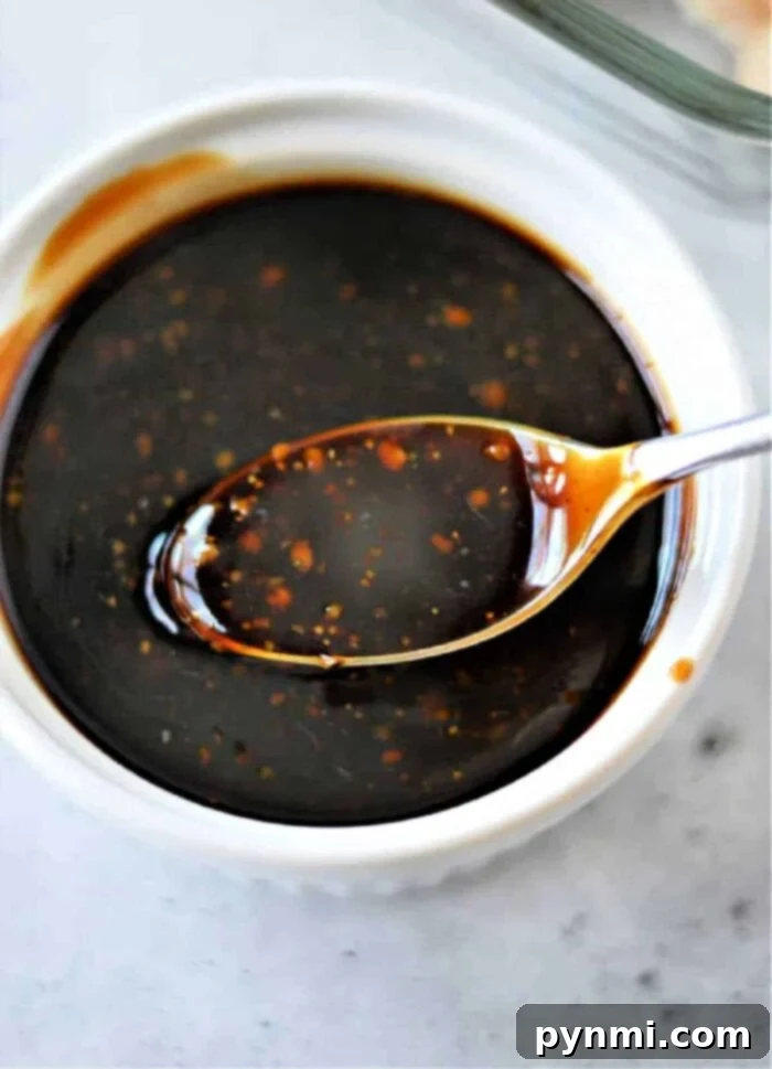 A spoon dipped in a bowl of dark teriyaki sauce with specks of seasoning, reminiscent of how to make homemade teriyaki sauce, rests on a white surface.