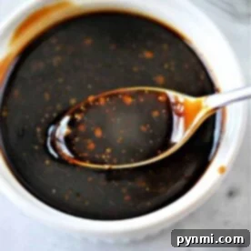 A spoon dipped in a bowl of dark teriyaki sauce with specks of seasoning, reminiscent of how to make homemade teriyaki sauce, rests on a white surface.
