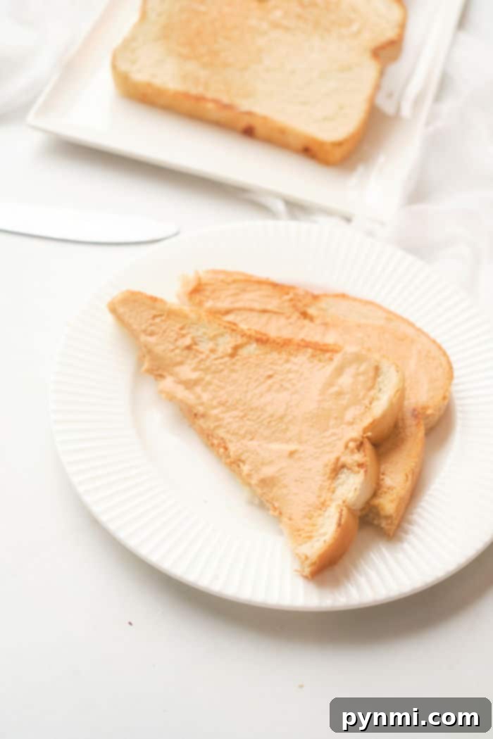 Sliced bread with peanut butter on a white plate, set on a table alongside a knife and more bread in the background, hints at the process of making toast in the oven for a warm, comforting start to your day.