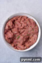 Ground venison mixed with herbs in a white bowl on a gray surface.