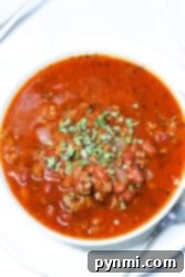 A bowl of Crockpot Venison Chili with minced meat, beans, and spices, garnished with chopped herbs, sits invitingly on a white plate accompanied by a spoon.