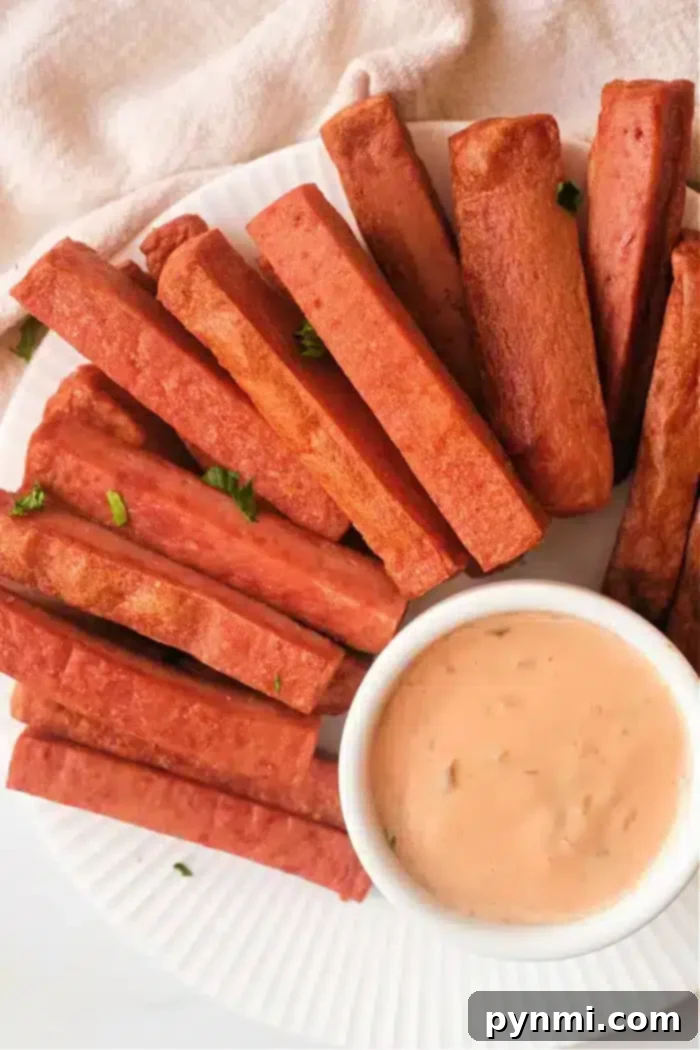 Crispy Air Fryer Spam Fries 2 A plate of rectangular fried Spam fries is arranged around a small dish of creamy dipping sauce on a white plate. Some parsley leaves are scattered for garnish. The background is a light-colored fabric.