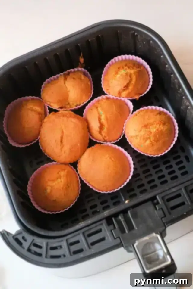 Effortless Air Fryer Cupcakes 4 Seven freshly baked air fryer cupcakes in pink silicone cups are arranged in the basket of a black air fryer. The cupcakes are golden-brown and appear to have a slightly cracked surface. The air fryer is positioned on a light-colored countertop.