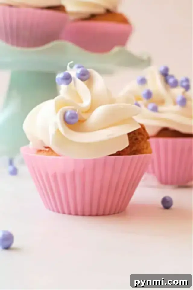 Effortless Air Fryer Cupcakes 8 A close-up of air fryer cupcakes in pink silicone wrappers, topped with white swirled frosting and decorated with small purple pearl-like sprinkles.