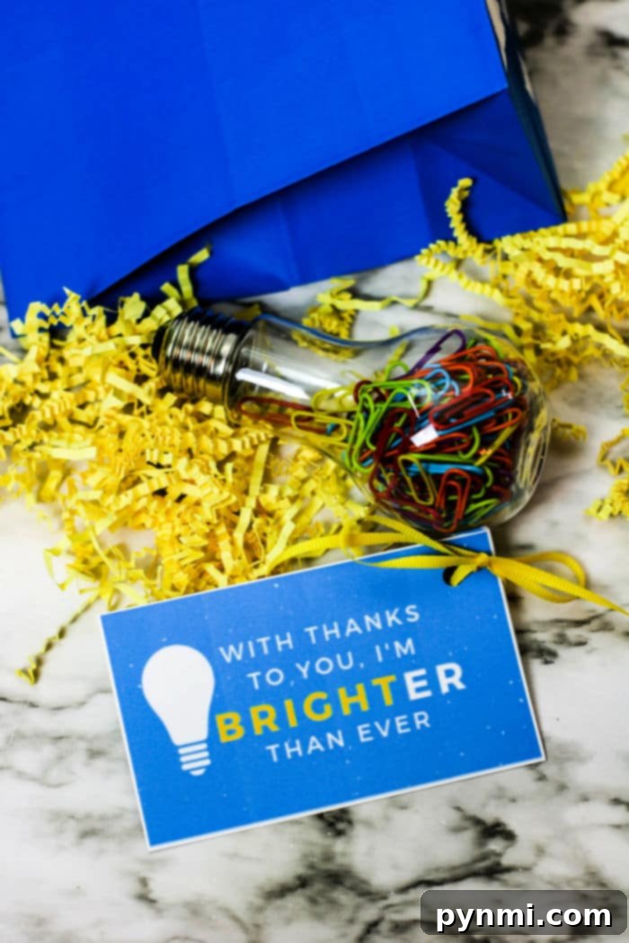 Free Printable Thank You Tags for Teachers 4 A blue gift bag on a marble surface contains yellow shredded paper and a clear bulb-shaped container filled with colorful paperclips. Alongside, a blue card with a lightbulb graphic reads, With thanks to you, I'm brighter than ever—perfect for attaching free printable teacher appreciation tags.