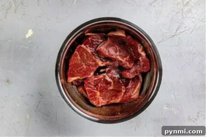 Ultimate Asian Steak Marinade 8 A metal bowl filled with raw pieces of beef, ideal for an Asian steak marinade, sits on a gray textured surface. The beef is marbled and visibly fresh. The bowl is the focal point of the image.