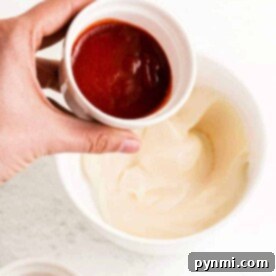 Easy Homemade Sriracha Mayo 6 A hand holds a small bowl of sriracha sauce above a bowl of mayonnaise, illustrating the process of making Sriracha mayo