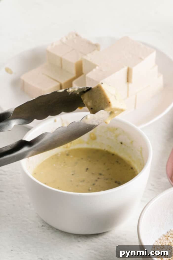 Golden Tofu Poppers 4 Tongs holding a tofu cube over a bowl of yellow sauce resemble the careful preparation of tofu nuggets, with more tofu blocks waiting on a plate in the background.