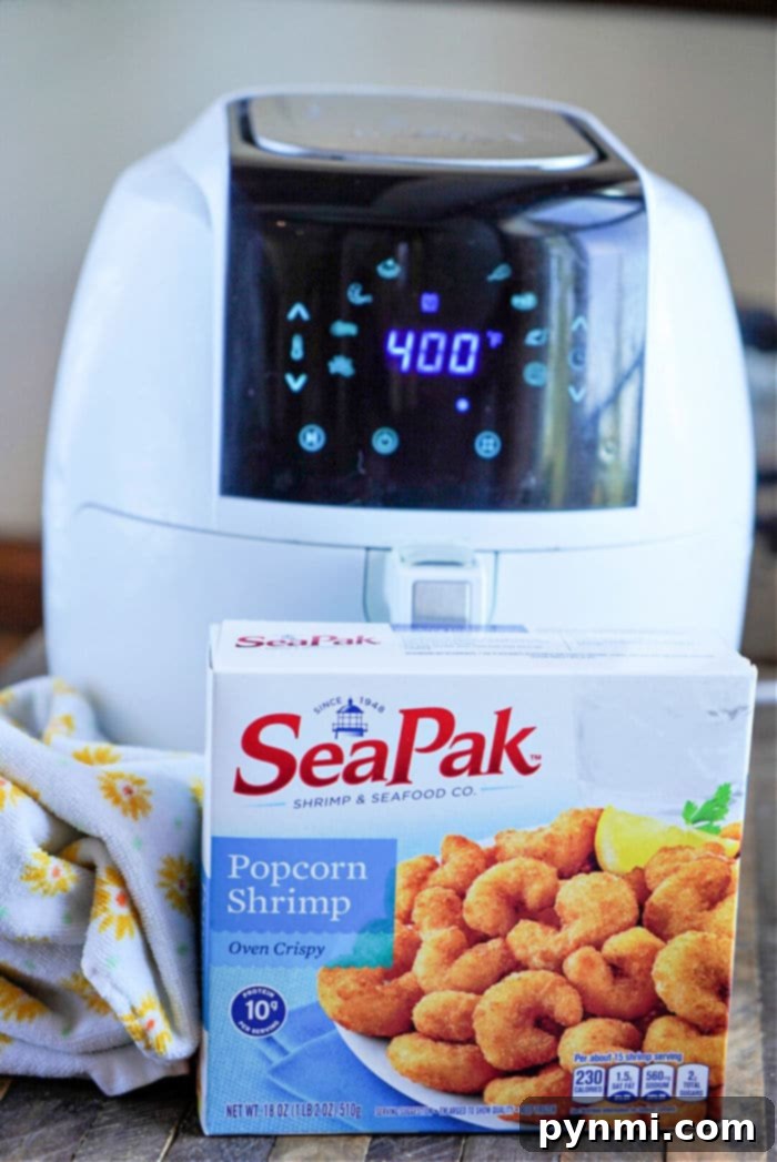 Crispy Air Fryer Popcorn Shrimp 4 A white air fryer, ideal for cooking frozen popcorn shrimp, displays 400 on its digital panel. In front of it is a box of SeaPak Popcorn Shrimp, labeled Oven Crispy, with a lemon wedge pictured on the packaging. A yellow and white patterned cloth is nearby.