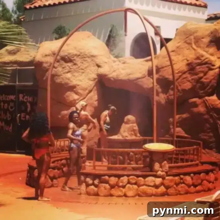 Experience the steamy allure of Glen Ivy Hot Springs' unique features, including tranquil pools and relaxation areas. People in swimsuits stand near a stone structure emitting steam outdoors at Glen Ivy in Corona. The background features large rocks and a building with a tiled roof. Some individuals are entering or exiting the steamy area, while a sign partially visible on the left reads "Welcome.