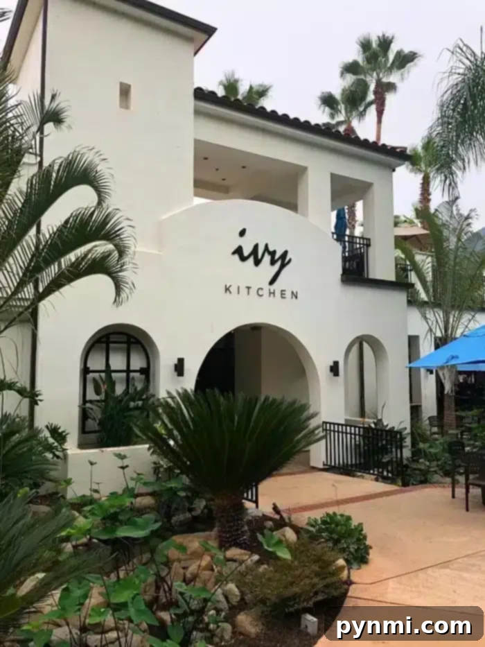 Indulge & Save: Glen Ivy Hot Springs Corona Specials 2 A white, two-story building with a sign reading "Ivy Kitchen" above the arched entrance is nestled in landscaped grounds featuring various plants and palm trees. A patio with a blue umbrella offers a cozy spot to enjoy Glen Ivy Corona deals.
