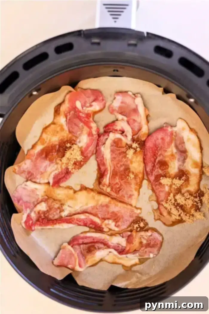 Crispy Candied Air Fryer Bacon 4 Slices of raw bacon, marbled with fat and kissed with maple syrup, are arranged on parchment paper inside an air fryer basket. The strips appear to be sprinkled with brown sugar, creating the perfect base for candied bacon.