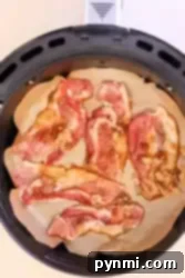 Slices of raw bacon, marbled with fat and kissed with maple syrup, are arranged on parchment paper inside an air fryer basket. The strips appear to be sprinkled with brown sugar, creating the perfect base for candied bacon.