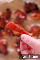 A hand holding a crispy piece of Air Fryer Maple Candied Bacon, with more maple-glazed bacon pieces blurred in the background.