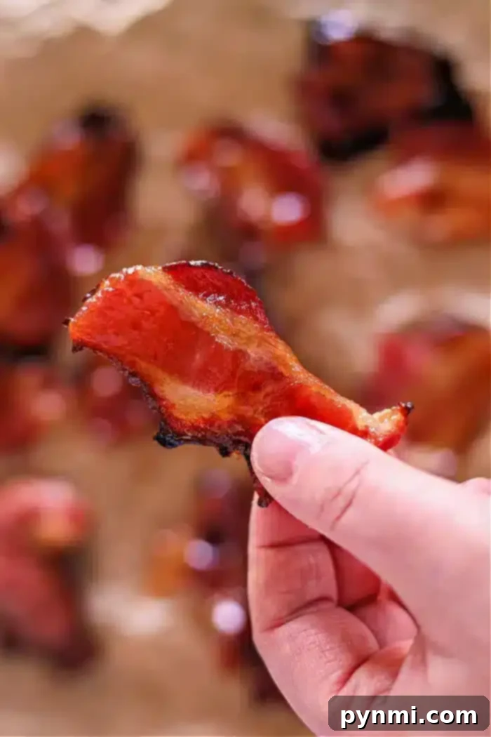 Crispy Candied Air Fryer Bacon 9 A hand holds a crispy piece of air fryer maple candied bacon with a glossy surface. Other pieces are blurred in the background, scattered on a light-colored surface.