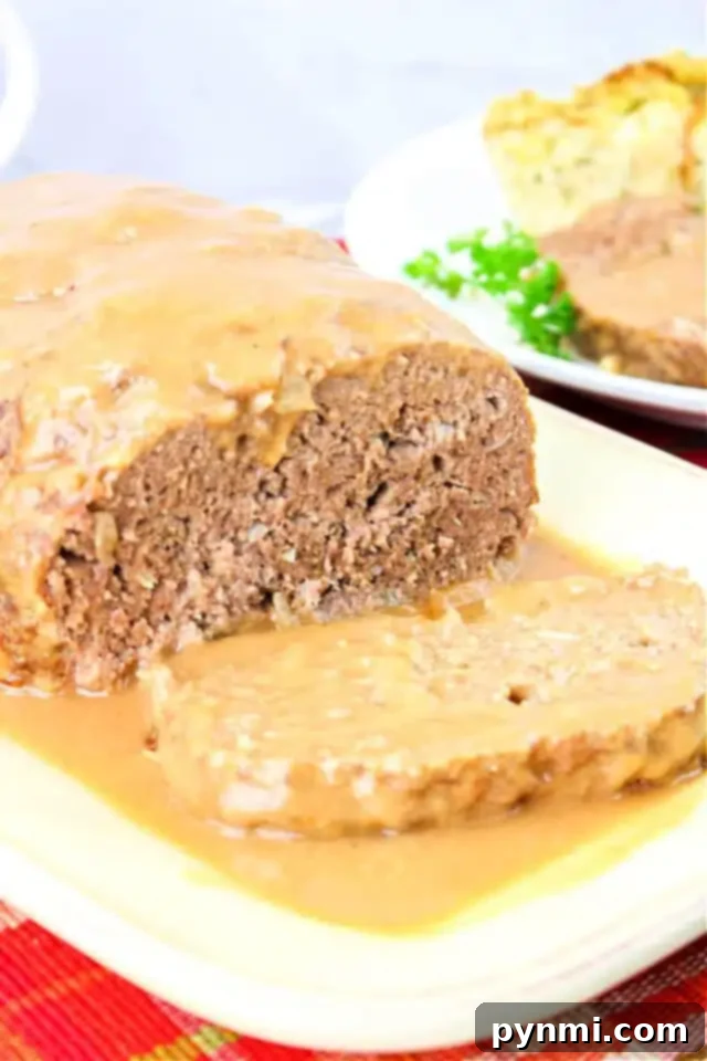 Foolproof Brown Gravy for Meatloaf 2 A loaf of meatloaf topped with rich brown gravy is served on a white plate. A slice has been cut from the front, revealing its texture. In the background, there is a plate with side dishes, including peas—a perfect scene for our savory brown gravy for the meatloaf recipe.