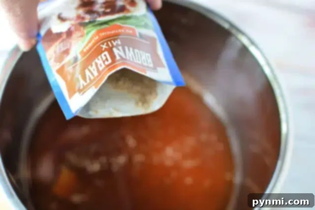 Foolproof Brown Gravy for Meatloaf 3 A hand is pouring a packet of brown gravy for meatloaf recipe mix into a metal pot filled with liquid. The contents are partially visible as they blend into the pot.