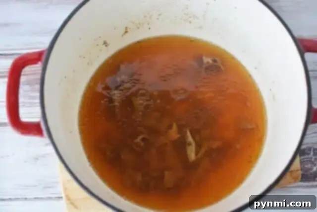 Foolproof Brown Gravy for Meatloaf 4 A red-handled pot containing simmering broth reminiscent of a brown gravy for meatloaf recipe with visible meat bones rests on a wooden surface. The rich, brown broth is slightly foamy at the edges.