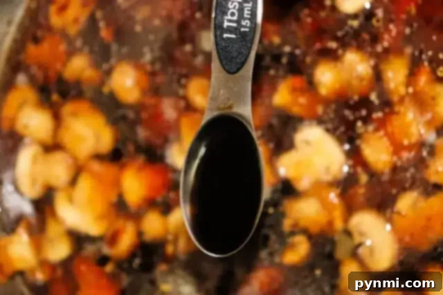 Foolproof Brown Gravy for Meatloaf 6 Close-up of a tablespoon labeled with 1 Tbsp, 15 mL, hovering above a pot of chickpeas, tomatoes, and spices. The spoon holds brown gravy, likely a rich seasoning for a meatloaf recipe.