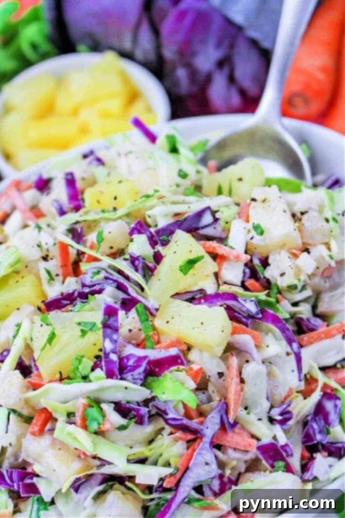 A vibrant bowl of Hawaiian coleslaw featuring vivid purple cabbage, crisp green cabbage, shredded carrots, juicy pineapple chunks, and a sprinkle of fresh herbs. A spoon rests alongside the bowl, inviting you to taste.