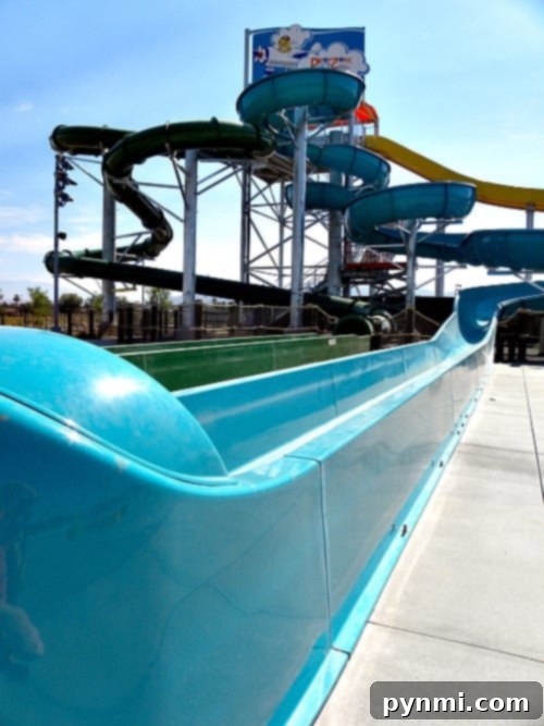 DropZone Perris Water Park: Is It Worth the Splash 5 A large water slide structure at Perris Water Park features multiple spiraling tubes of green and yellow. The blue slide leads to a splashdown area on a concrete surface. A sign with a cartoon figure is visible at the top of the structure. The sky is clear and sunny.