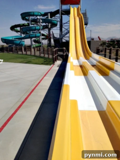 DropZone Perris Water Park: Is It Worth the Splash 6 A view of Perris Water Park's slide area features multiple yellow and white slides side by side. In the background, a spiraling green and blue water slide structure is visible. The area around the slides is paved, and there is a fence separating the slides from the grass.