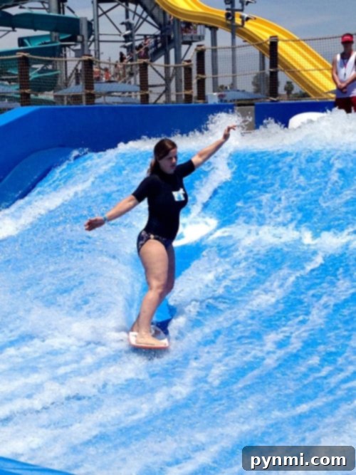 DropZone Perris Water Park: Is It Worth the Splash 8 A person in a black wetsuit rides a wave on a bodyboard at Perris Water Park, maintaining balance with arms outstretched. A yellow water slide is visible in the background, along with several other people looking on.
