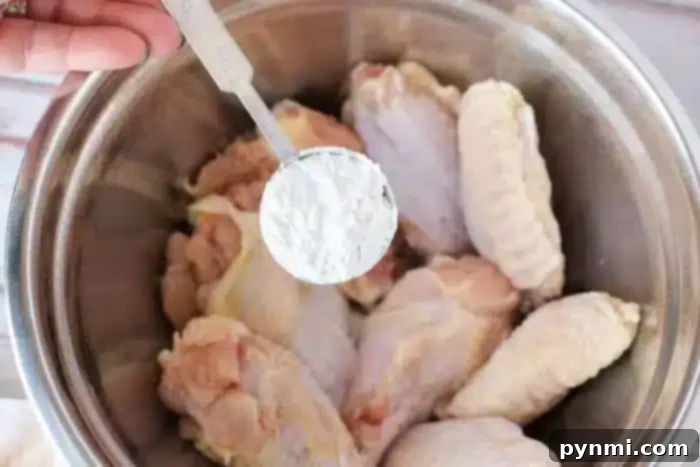 Crispy Air Fryer Wings Baking Powder Secret 5 A hand holding a tablespoon of white powder, likely baking powder, hovers over a metal bowl filled with raw chicken wings, ready to be tossed and cooked into crispy air fryer chicken wings.