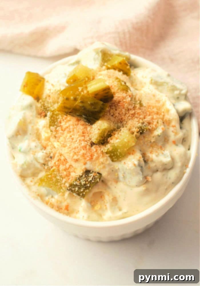 A serving bowl filled with creamy ranch pickle dip, garnished with chopped fresh dill and mini gherkins, surrounded by a variety of dippers like potato chips and vegetable sticks.
