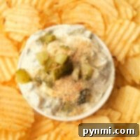 A bowl of Fried Pickle and Ranch Dip crowned with pickles and seasoning, accompanied by ripple-cut potato chips on a white plate, showcasing a delightful appetizer.