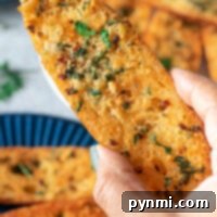 A hand holding a slice of golden garlic bread, freshly made from scratch and garnished with herbs, with more slices on a blue plate in the background.