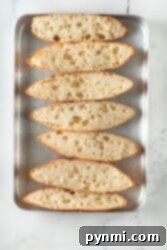 Sliced ciabatta bread with an airy texture on a metal baking sheet, buttered and ready for baking into garlic bread.