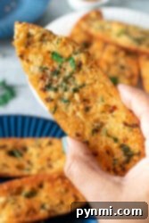 A hand holding a slice of golden garlic bread, freshly made and garnished with herbs, with more slices on a blue plate.