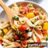 A bowl of roasted vegetable pasta salad with penne, tomatoes, olives, zucchini, and bell peppers, served with a wooden spoon.