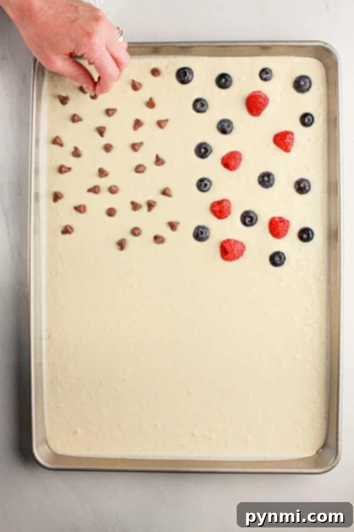A hand decorates oven baked pancakes with pancake mix in a tray, adding chocolate chips, blueberries, and strawberries in a flag pattern.