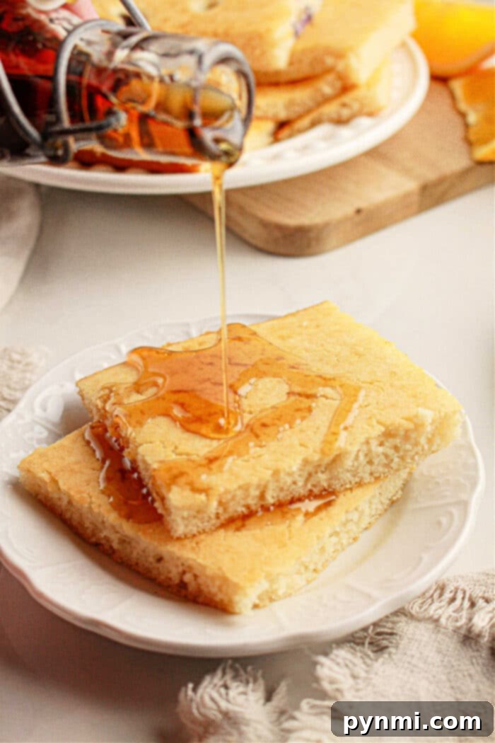 Pouring syrup over two stacked pieces of cornbread on a white plate, with more oven baked pancakes with pancake mix in the background.