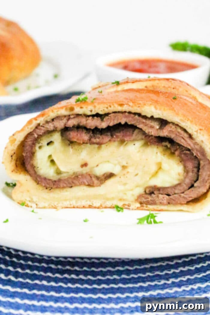 A sliced Philly Cheesesteak Stromboli with layers of beef and cheese on a white plate, sauce in the background.