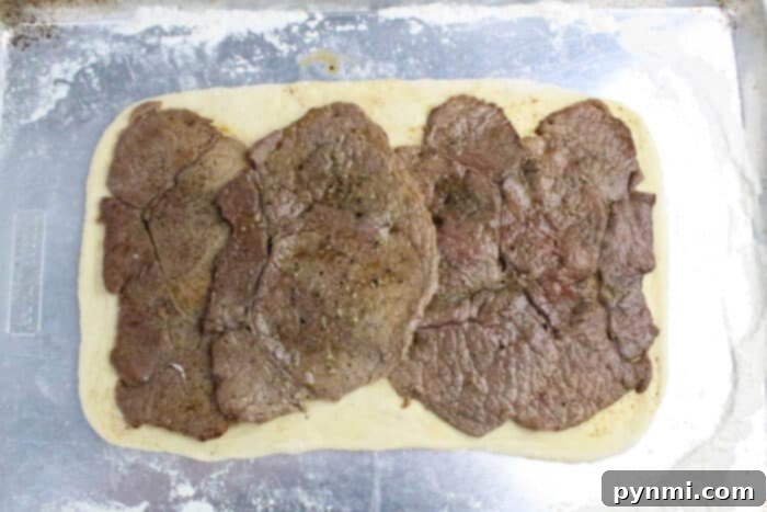 Rectangular dough topped with several overlapping slices of cooked beef, ready to be transformed into a delicious Philly Cheesesteak Stromboli on a floured baking sheet.