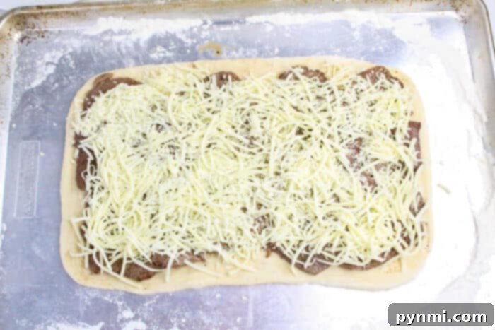 Rectangular pizza dough topped with meat slices and shredded cheese on a floured baking sheet, perfect for making Philly Cheesesteak Stromboli.