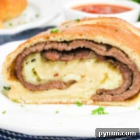 A sliced Philly Cheesesteak Stromboli with layers of beef and cheese on a white plate, sauce in the background.