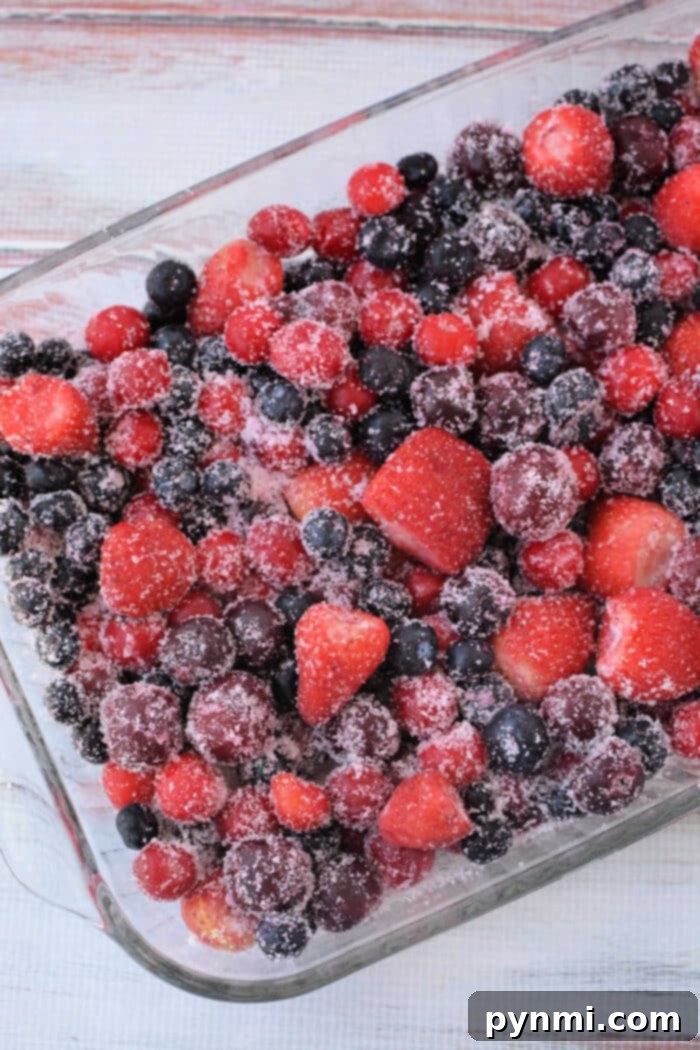 Cake Mix Berry Cobbler 4 A clear glass baking dish brimming with a colorful mix of frozen blueberries, cherries, and strawberries, generously coated with granulated sugar, ready for the next step in preparing a delicious Berry Cobbler with Cake Mix.