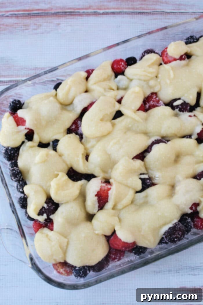 Cake Mix Berry Cobbler 5 An unbaked Triple Berry Cobbler with Cake Mix in a glass baking dish, featuring a vibrant layer of mixed berries at the bottom, topped with dollops of the rich cake mix and butter batter, ready to be baked into a comforting dessert.