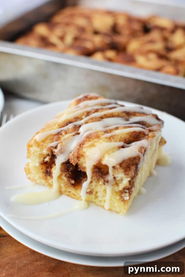 Semi-Homemade Cinnamon Roll Cake 2 A generous slice of moist cinnamon roll cake made from a cake mix, beautifully drizzled with creamy white icing, rests on a pristine white plate. In the soft-focused background, the rest of the freshly baked cake sits invitingly in its pan.
