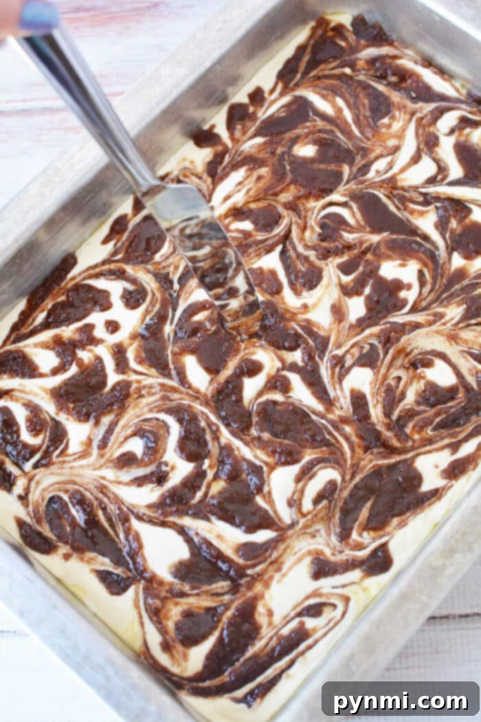 Semi-Homemade Cinnamon Roll Cake 4 A skillful hand expertly swirls a rich chocolate mixture into a light cake batter within a metal baking pan using a knife, creating a beautiful marbled effect. This technique is perfect for achieving the signature swirl in a homemade Cinnamon Roll Cake, promising layers of flavor.