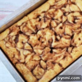 A Cinnamon Roll Cake with a beautiful cinnamon swirl pattern, baked in a rectangular metal pan using an easy cake mix, rests on a light wooden surface.