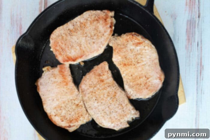 Apple-Glazed Baked Pork Chops 4 Four perfectly seared and seasoned pork chops cooking in a black cast iron skillet, surrounded by softening apple slices, on a light wooden surface, illustrating the initial cooking phase of baked pork chops with apples.