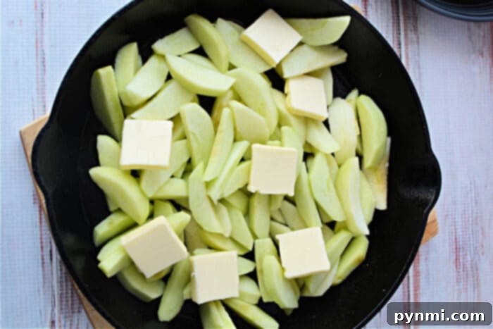 Apple-Glazed Baked Pork Chops 5 Neatly sliced Granny Smith apples topped with pats of rich butter, ready to be cooked down in a black skillet on a light wooden surface, highlighting the apple preparation for pork chops with apples.