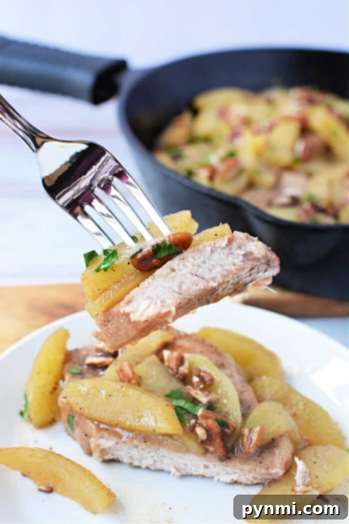 Apple-Glazed Baked Pork Chops 6 A close-up of a fork holding a succulent bite of baked pork chop crowned with tender cooked apples and crunchy pecans, above a beautifully plated serving with more apples and a cast iron skillet in the background. A perfect display of the finished dish.